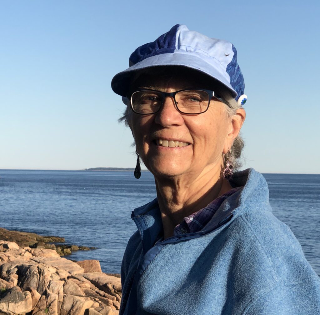 I am an older woman with pale peach skin and white hair. In this picture I wear a light blue fleece jacket and a cap with light blue and dark blue sections. I wear glasses and dangling earrings. I look drectly at the viewer with a big smile. Behind me is blue sky and deeper blue ocean with pinkish granite boulders on one side.