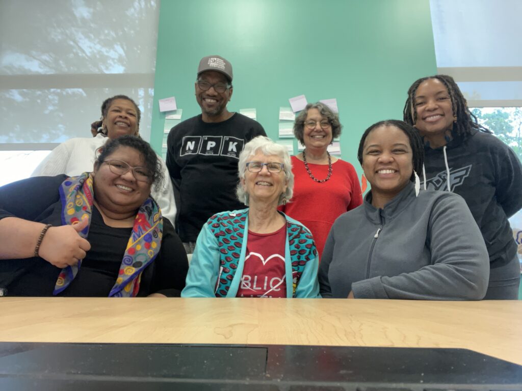 A group of people sitting at a table. All are smiling brightly. Jeanie sits in the middle. Four women and one man have brown skin while Jeanie and another woman have lighter peach skin tones. One woman is giving a thumbs-up sign.