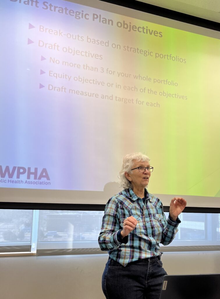 I am facilitating strategic planning with Wyoming Public Health Association. Wearing a turquoise plaid blouse, I stand in front of a screen where a Power Point slide shows the steps in drafting strategic planning objectives.