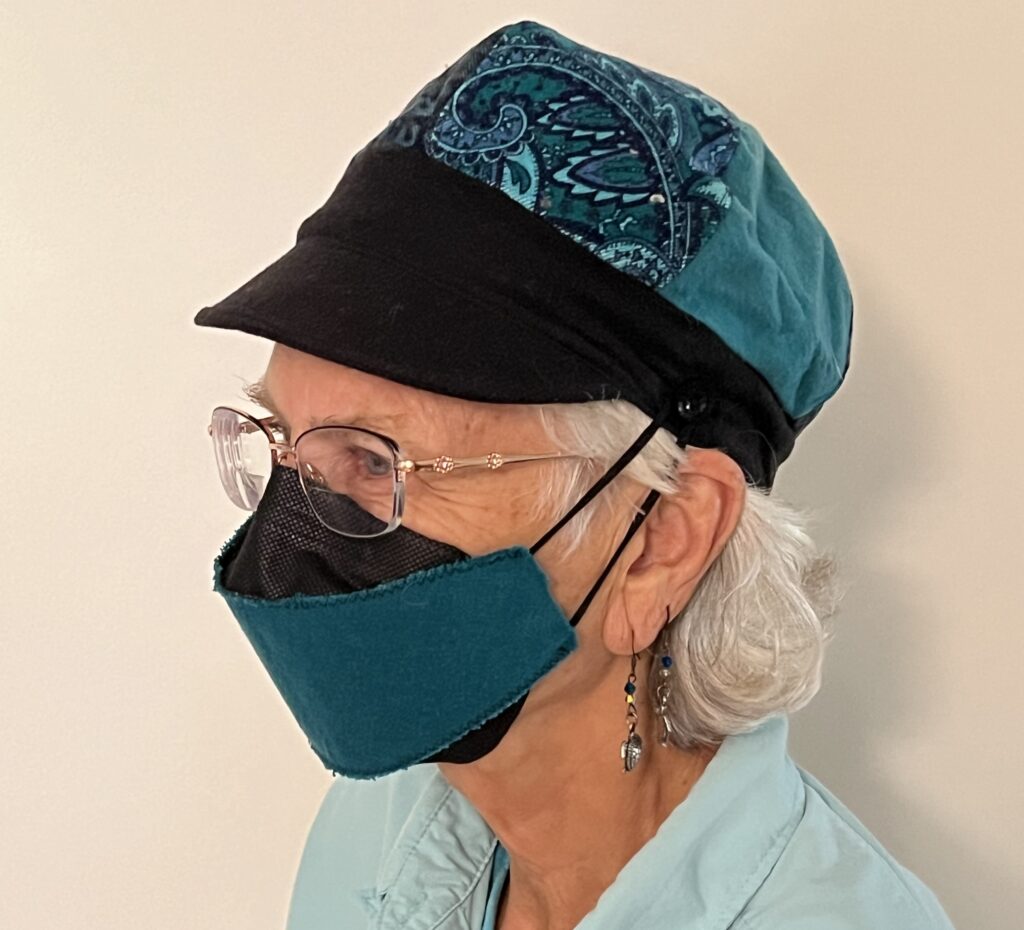 I like to make N95 masks a little fancier and make them match what I wear. I also like to make and wear hats. In this picture I wear a newsy-style hat with a short bill. The hat is patterned teal and turquoise. The hat has a black brim and bill. The black N95 mask has a piece of teal fabric sewn on the from so that it matches the hat..